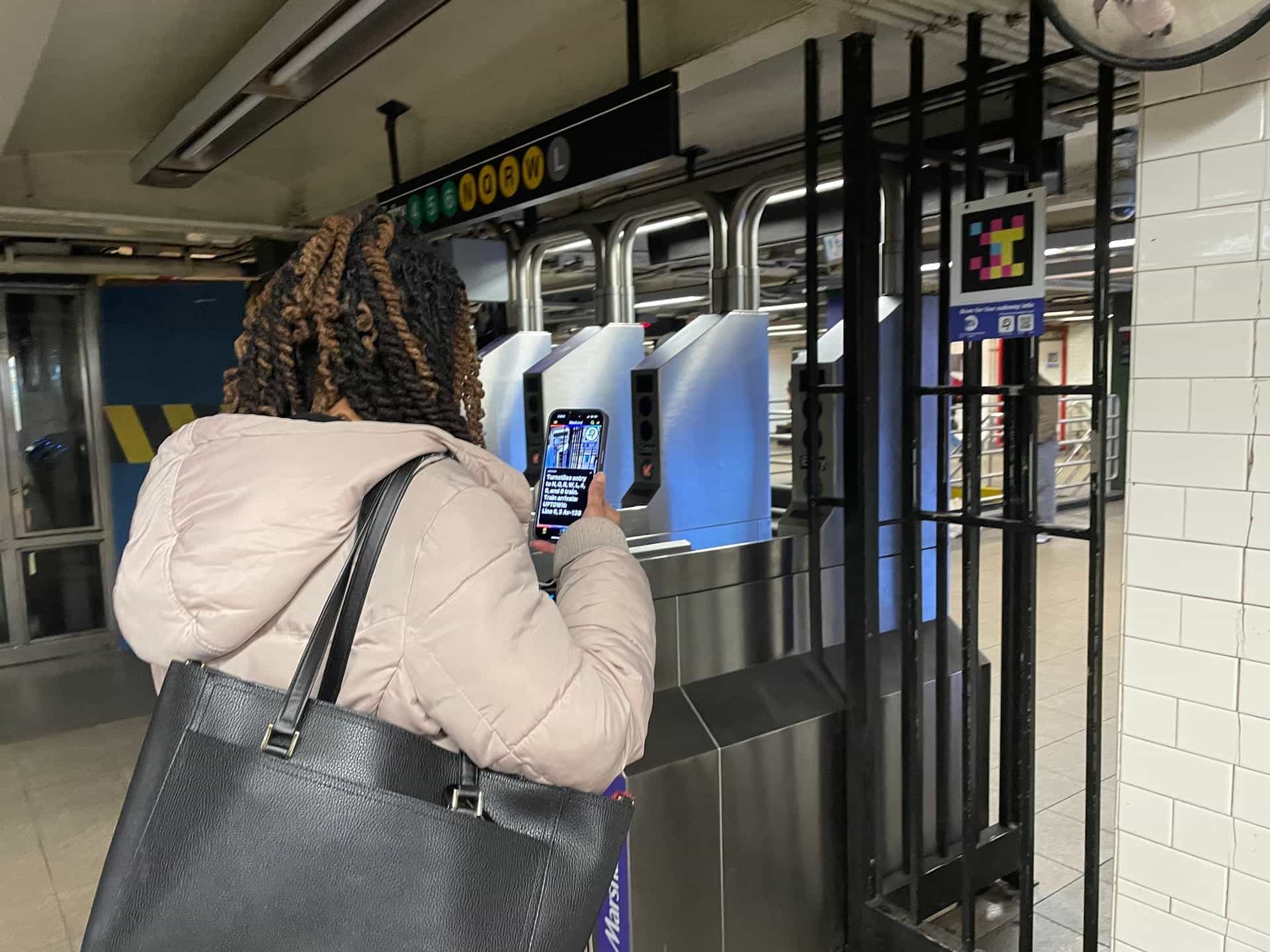 Pasajera en una estación de metro apuntando su teléfono hacia un código NaviLens ubicado junto a los torniquetes